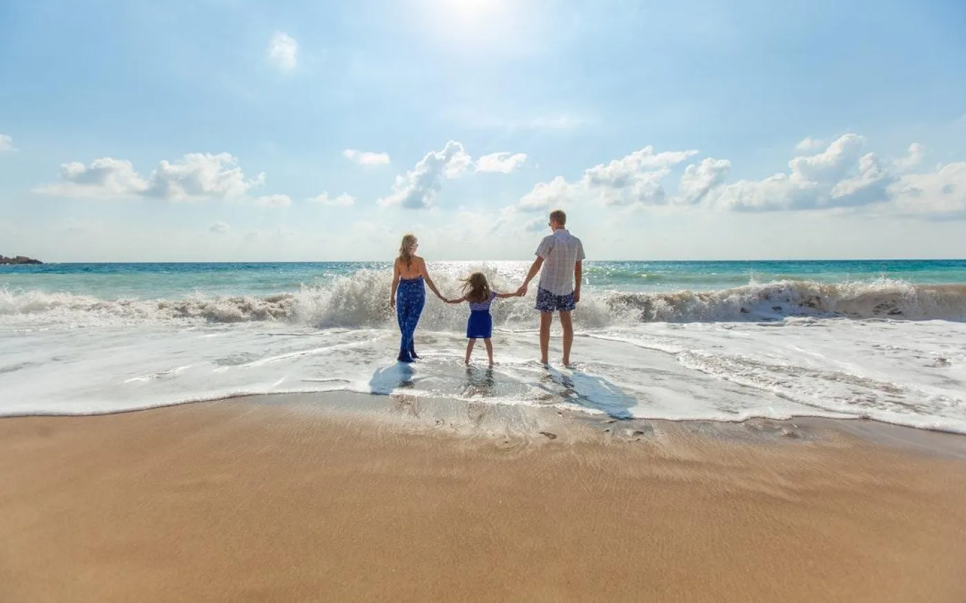 A family stands hand in hand on the beach, with waves gently lapping at their feet under a clear blue sky.