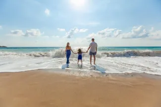 A family stands hand in hand on the beach, with waves gently lapping at their feet under a clear blue sky.