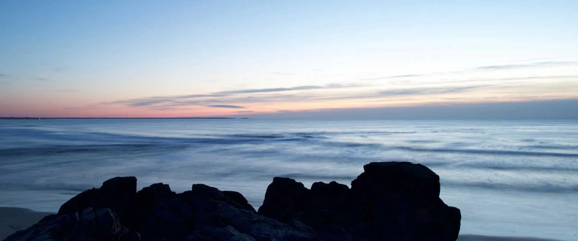A rocky beach at dusk, with gentle waves lapping against the shore under a colorful sky.