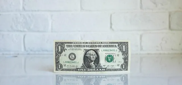 A one dollar bill placed on a table in front of a textured white brick wall.