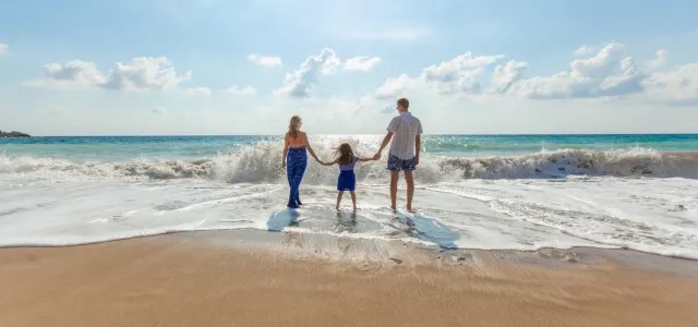 A family stands hand in hand on the beach, with waves gently lapping at their feet under a clear blue sky.